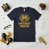 Dark navy t-shirt with bold gold text reading "60 BIRTHDAY SQUAD" in a vintage collegiate style, featuring a shield and
