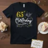 Black t-shirt with "65th Birthday Crew" text in white and yellow, featuring festive graphics like confetti and party poppers
