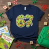 A navy t-shirt featuring the number 67 formed by two cute green snakes with orange spots, set against a wooden