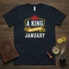 A King Was Born In January text design with a crown and shield graphic on a black t-shirt