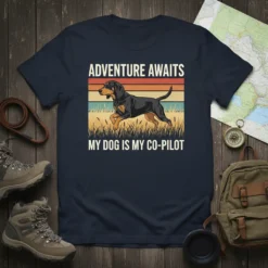 Graphic t-shirt with white text "ADVENTURE AWAITS MY DOG IS MY CO-PILOT" and a running dog illustration on a sunset