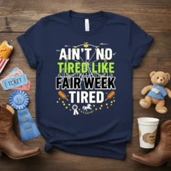 Navy t-shirt with "Ain't No Tired Like Fair Week Tired" in bold white and green lettering, featuring carnival graphics like