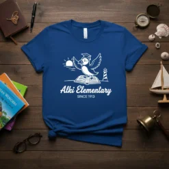 Alki Elementary Since 1913 t-shirt featuring a white puffin mascot, graduation cap, open book, sun, and lighthouse graphic