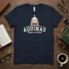 Navy t-shirt featuring "AQUINAS SCHOLA OPTIMA" text and a building graphic, representing academic pride