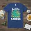 Auntie Saurus design on a blue t-shirt featuring a green T-Rex flexing its arm among tropical leaves