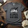 T-shirt with "BE CAREFUL OR YOU'LL END UP IN MY BOOK" quote and open book graphic with quill