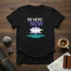 Black t-shirt with "BE HERE NOW" text above a white and purple lotus flower in water, primary keyword included