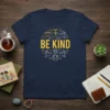 Navy blue t-shirt with "BE KIND" text in yellow, surrounded by doves, hands making a heart, and globes