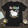 Black t-shirt with "Be Kind" text in pastel colors, featuring a cute unicorn sheep surrounded by flowers and a rainbow