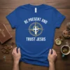 Christian t-shirt with white text 'BE PRESENT AND TRUST JESUS' and a cross inside a compass graphic