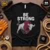 Black t-shirt with bold white text "BE STRONG" above a graphic of a powerful bull in maroon and grey