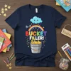 Be A Bucket Filler! text with a rainbow, cloud, and bucket filled with stars on a navy blue t-shirt