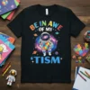 Black t-shirt with "Be In Awe Of My 'Tism" text and an astronaut holding a puzzle heart, set against a