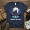 Bigfoot Believes text on a navy t-shirt with a graphic of Bigfoot standing in water under a full moon