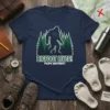 Navy t-shirt with white and green Bigfoot silhouette in a forest, text reads "BIGFOOT LIVES! PACIFIC NORTHWEST"