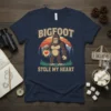 Bigfoot Stole My Heart T-Shirt design featuring a cartoon Bigfoot holding a lantern with a heart, set against a sunset