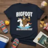 Bigfoot riding a unicorn float with text "BIGFOOT ON A UNICORN FLOAT" on a navy t-shirt