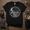 Black Cat Social Club text design with circular graphic featuring cats, moon, bats, and flowers on a black t-shirt