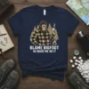 Bigfoot in plaid shirt holding marshmallow saying "Blame Bigfoot He Made Me Do It" on a navy t-shirt design