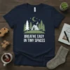 Breathe Easy In Tiny Spaces t-shirt with a graphic of a small cabin in a forest under a crescent moon