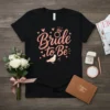 Black t-shirt with "Bride to Be" text in coral script, adorned with hearts, a diamond ring, and a dove graphic