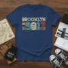 Navy blue t-shirt with "BROOKLYN 9917" in colorful vintage font and a cityscape graphic below