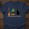 Broomstick, cauldron with green brew, and black cat illustration on a navy blue t-shirt, with text "Broomstick Brew Black Cat"