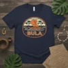 Bula text with a sailboat, palm trees, and ocean waves graphic on a navy t-shirt