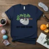 Navy blue t-shirt with a cute grey sloth hanging from a tree branch, text reads "Busy Doing Nothing"