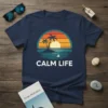 Calm Life text with a tropical sunset, island, palm trees, and sailboat graphic on a navy blue t-shirt