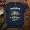 Navy blue t-shirt featuring the text "CAMPING DAD LIKE A REGULAR DAD BUT Cooler" with a vintage camper, campfire, and