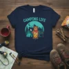 Camping Life t-shirt design with a cartoon bear roasting a marshmallow by a campfire, surrounded by trees and mountains