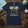 Black cat with "CAT LIFE EXPLORE DISCOVER PLAY" text and celestial graphics on a navy t-shirt