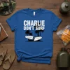 Blue t-shirt with white text CHARLIE DON'T SURF and a graphic of a broken surfboard on a beach with palm