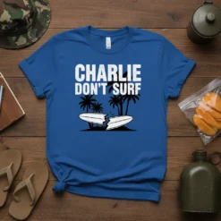 Blue t-shirt with white text CHARLIE DON'T SURF and a graphic of a broken surfboard on a beach with palm