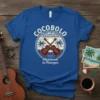Royal blue t-shirt featuring a circular design with "COCOBOLO Ukuleles Handmade in Nicaragua" text, two crossed ukuleles, palm trees, and