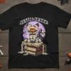 Black t-shirt featuring a cartoon mummy cat sitting on books with "Coffin Creed" text above, set against a purple moon