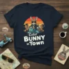 Coolest Bunny in Town graphic tee with a rabbit riding a scooter against a sunset city backdrop