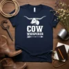 Navy blue t-shirt with white text "Cow Whisperer" and a longhorn cow graphic, featuring an arrow detail