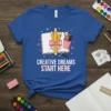 Creative Dreams Start Here quote on a blue t-shirt with pencils, a notebook, and art supplies graphic