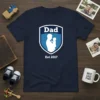 Dad Est 2017 t-shirt design featuring a shield with a father holding a baby silhouette and the year 2017