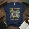 T-shirt design with text "DEFEND YOUR LOCAL PUBLIC LIBRARY" featuring a cat in knight armor riding a greyhound