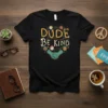 Black t-shirt with "Dude Be Kind" text design, featuring diverse illustrated faces and shaking hands in a circular pattern