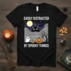 Black t-shirt with white text "Easily Distracted By Spooky Things" and a Halloween graphic of a black cat, ghost, pumpkin