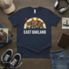 East Oakland text overlay on a navy t-shirt featuring a California bear walking across a stylized cityscape under a sunburst