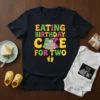 EATING BIRTHDAY CAKE FOR TWO text with birthday cake graphic on a black t-shirt, baby feet icon below