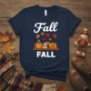 Navy t-shirt with "Fall FALL" text, featuring a coffee cup, stack of books, and pumpkin graphic, surrounded by autumn leaves