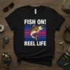 "Fish On! Reel Life" text with a jumping fish graphic on a black t-shirt, ideal for fishing enthusiasts
