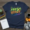 Navy blue t-shirt with "Fresh Start" in bold green and orange lettering, featuring a yellow arrow graphic and colorful confetti