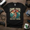 T-shirt with a cartoon T-Rex wearing VR goggles, playing video games, and the text "GAME OVER PRESS START TO CONTINUE"